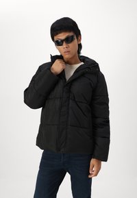 Black puffer jacket with a zip front, hood, and stitched panels. Worn with a light-colored shirt and dark jeans, paired with black sunglasses.