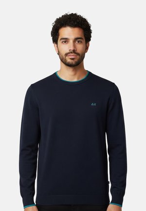 Uomo che indossa un maglione a maniche lunghe blu navy con bordi verde acqua al colletto e ai polsi, in piedi davanti a uno sfondo bianco semplice.
