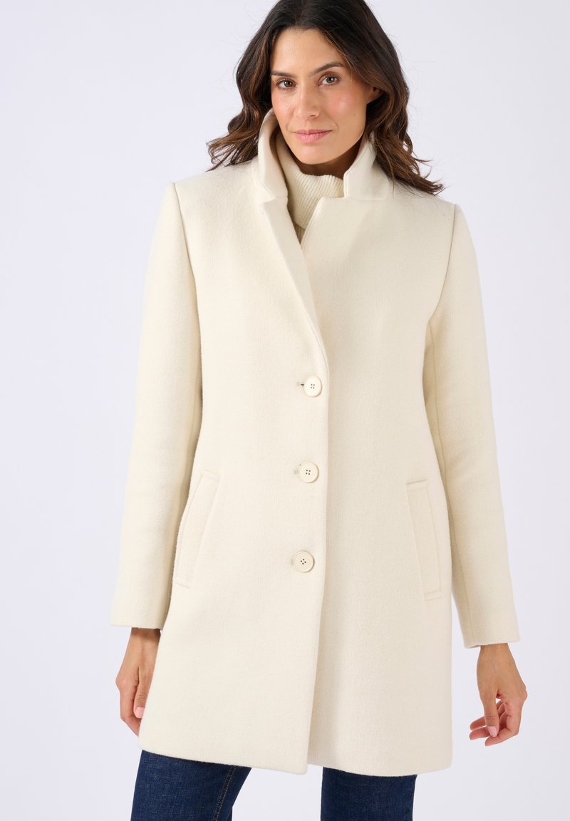 Cappotto bianco in lana con colletto alto, quattro grandi bottoni, tasche laterali e un taglio dritto. Texture liscia, design classico, accenti minimi.