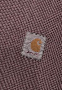 Vaflinės tekstūros audinys švelnaus violetinio atspalvio, su kvadratine Carhartt logotipo etikete, kurioje yra stilizuotas geltonas 'C' ir raudoni užrašai.