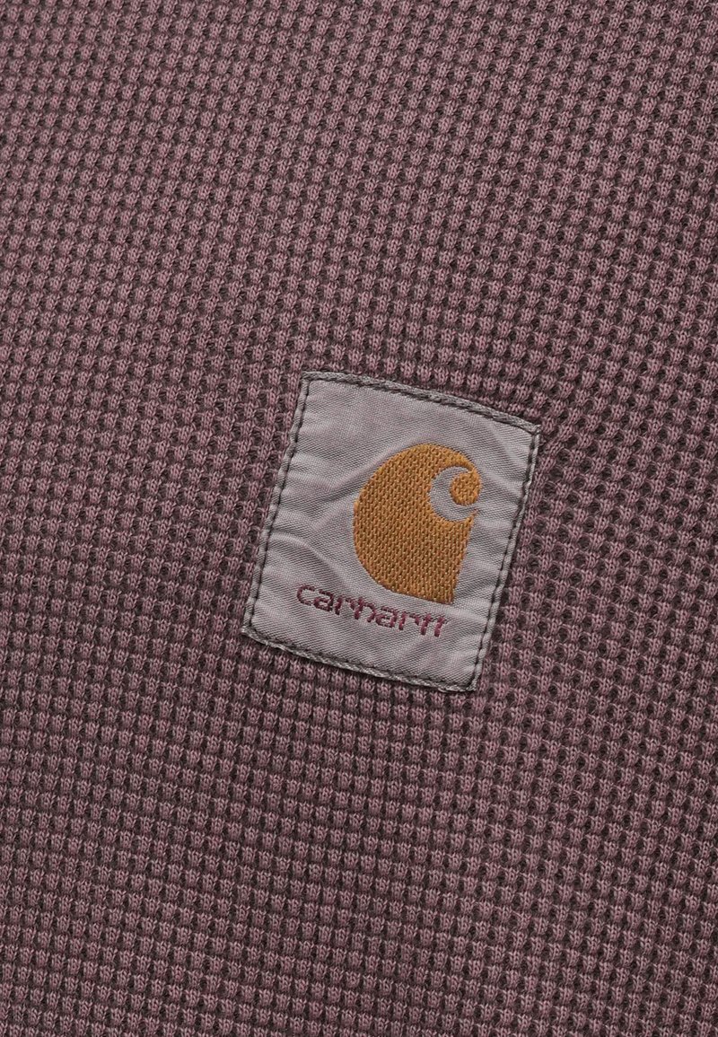 Vaffelstruktureret stof i en dæmpet lilla tone med et firkantet Carhartt-logo i form af en gul 'C' med stiliseret design og røde bogstaver.