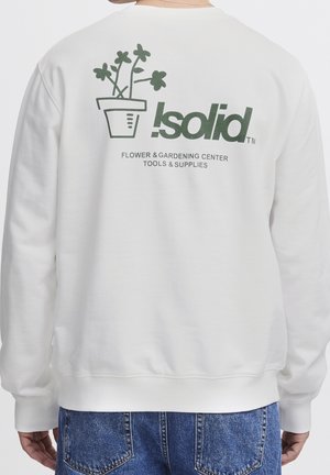 Osoba nosząca biały sweter z zieloną grafiką kwiatka w doniczce i napisem „Isolid Flower & Gardening Center Tools & Supplies” na plecach.