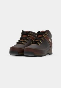 Botas de senderismo de cuero marrón con cordones negros, ojales de metal, cuellos acolchados y suelas de tracción resistentes. Detalle de costura reforzada.
