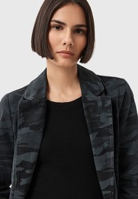 Blazer nero camouflage con rever a punta, maniche lunghe e tessuto leggermente strutturato, abbinato a un top nero con scollo rotondo sottostante.