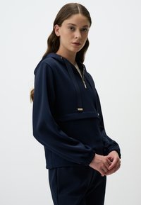 Felpa blu navy con zip metà lunghezza, dettagli dorati, cappuccio con cordino ed elastici ai polsi. Realizzata in un materiale liscio e morbido.