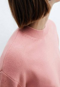 Personne aux cheveux bruns courts portant un pull en tricot rose clair avec un col côtelé, photographiée de l'épaule vers le haut, légèrement tournée sur le côté.