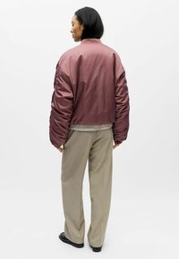 Burgunderfarget bomberjakke med blank finish, ribbestrikket kant og voluminøse ermer, kombinert med lysegrå plissert bukse.