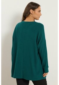 Pull teal surdimensionné avec manches longues, fabriqué en tissu doux. Il présente un ourlet arrondi et des détails de couture au dos. Aucun motif ni matériel visible.