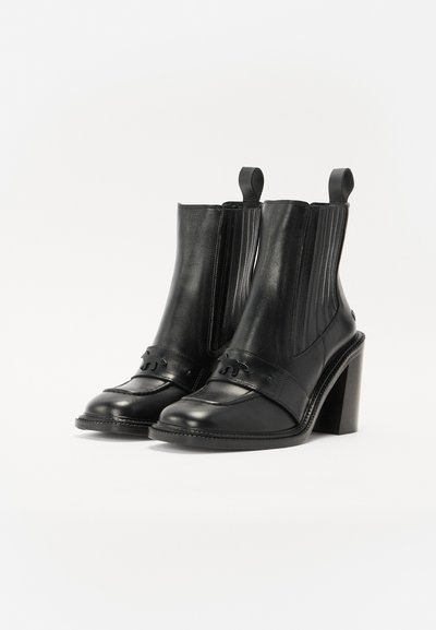 Botines de piel negra que cuentan con un tacón bloque, paneles laterales elásticos y una correa decorativa con un pequeño logotipo. Textura suave, diseño minimalista.