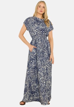 Vrouw draagt een maxi-jurk met een marineblauw-wit abstract patroon, korte mouwen, elastische taille en zakken, staand met één hand in de zak.