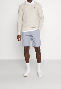Béžový pletený svetr s jemným texturovaným vzorem, doplněný o světle modré šortky. Bílí tenisky a ponožky dotvářejí outfit.