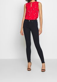 Blouse sans manches rouge avec un col en trou de serrure, imprimé floral et bords froncés, associée avec un jeans skinny noir et des talons noirs à bride de cheville.