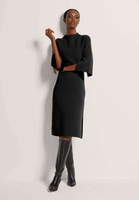Schwarzes, figurnahes Strickkleid mit dreiviertellangen Glockenärmeln, seitlichen Schlitzen und hohem Kragen, kombiniert mit eleganten knieh hohen schwarzen Stiefeln.