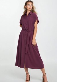 Fioletowa sukienka zapinana na guziki do kolan z krótkimi rękawami, z paskiem w talii i rozcięciem po boku. Gładki materiał o luźnym kroju.