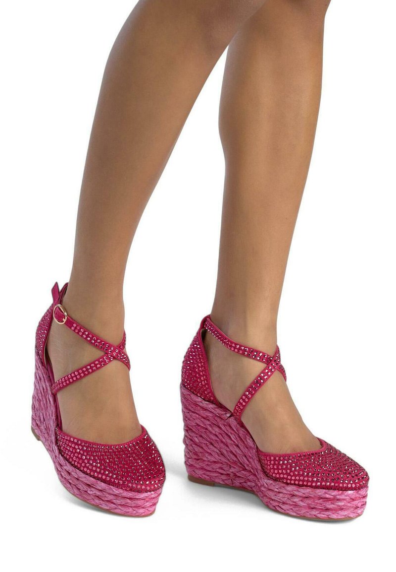 Des chaussures à talons compensés roses avec une base tissée, ornées d'accents en strass scintillants et de bretelles croisées, présentant une finition lisse et texturée.