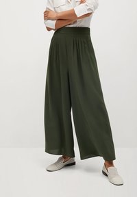 Pantalons larges, verts olive, avec des plis à la taille, fabriqués en tissu lisse. Assortis à des chaussures légères et texturées à enfiler.