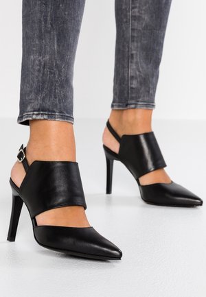 Czarne skórzane buty na wysokim obcasie typu slingback z ostro zakończonymi noskami, noszone z wyblakłymi czarnymi obcisłymi jeansami.