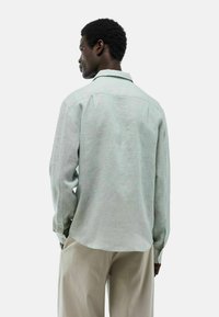 Chemise en lin vert clair à manches longues, avec des poignets à boutons, un détail de pattes de boutonnage au dos, et une coupe décontractée. Portée avec un pantalon beige, vue de dos.