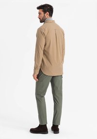 Khaki overhemd met knopen en een rugplooien, gecombineerd met olijfgroene broek en bruine suède enkelboots, gezien vanuit achteren.