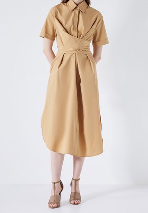 Femme portant une robe midi beige à manches courtes avec col, détail ceinture portefeuille et sandales à talons hauts ouvertes assorties beige.