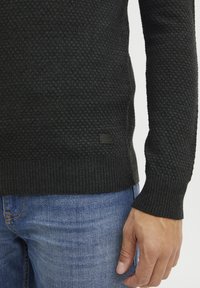 Svart texturerad tröja med ett honungskakemönster, ribbade ärmslut och nederkant; har en liten läderetikett nära nedre vänstra delen. Bärs med blåjeans.