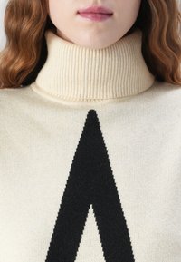 Kerman neulepusero ribbed-kudoksella, jossa on suuri musta kolmion muotoinen kuvio näkyvästi esillä edessä.
