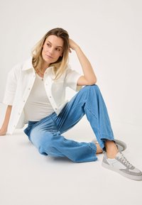 Cămașă albă cu nasturi, top gri deschis fără mânecă, pantaloni din denim albastru, adidași gri și albaștri; ținută casual cu o croială lejeră și țesătură texturată.