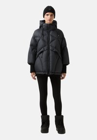 Person iført en sort dunjakke med hætte, sorte leggings, sorte vinterstøvler og en sort strikket hue, stående mod en ensfarvet baggrund.