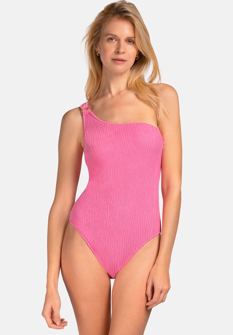 Blondynka w jednoczęściowym, różowym kostiumie kąpielowym na jedno ramię, z detalem w postaci pierścienia na pasku, stoi na tle jednolitego białego tła.