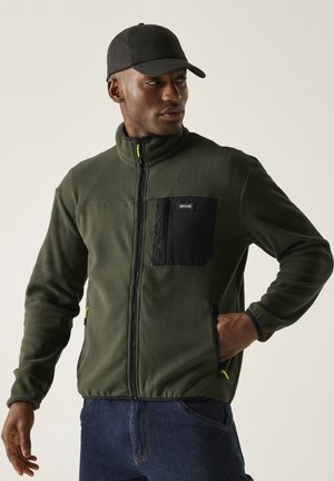 Homme portant une veste polaire vert foncé avec une poche poitrine noire et une casquette noire, debout les mains dans les poches devant un fond uni.