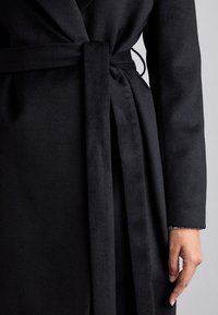 Manteau en laine noir au design enroulé, avec une ceinture nouée et des poignets texturés. Tissu lisse, détails de couture minimaux.