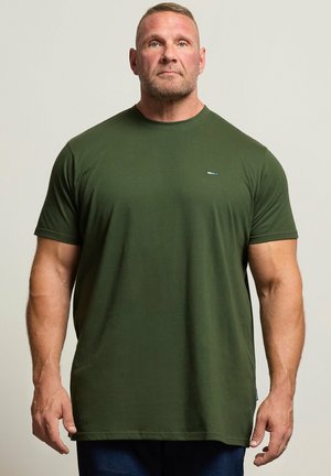 Muskuløs mand med kort hår står med fronten fremad, iført en ensfarvet mørkegrøn t-shirt og mørke bukser mod en neutral baggrund.