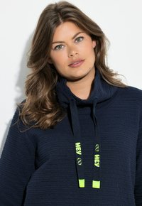 Jersey de punto azul marino con un patrón texturizado, cuello alto y cordones ajustables amarillo neón contrastantes con la marca "HEY".