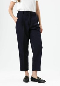 Marineblauwe hoge taillebroek met plooien, zijzakken en een rechte pijp. De stof oogt glad; gecombineerd met zwarte loafers.