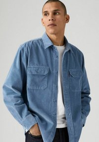 Camicia in corduroy blu chiaro con due tasche sul petto, chiusura a bottoni e maniche lunghe. Texture liscia con un evidente motivo a coste.