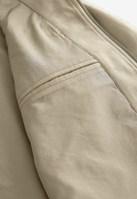 Veste beige avec une texture lisse et mate. Présente une fermeture éclair fine, une poche sur la couture latérale et des accents doux semblables à du daim le long du col.