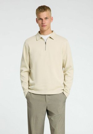 Beige pullover med lynlås krave, tekstureret stof og lange ærmer. Båret over en hvid skjorte, kombineret med lysegrå bukser.