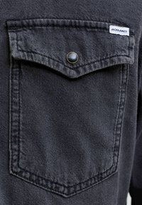 Közeli kép egy fekete farmering zsebéből, amelyen gombos fedő található, és a jobb felső sarokban egy kis fehér "JACK & JONES" címke látható.