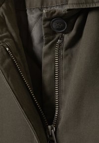 Detail einer olivgrünen Stoffjacke mit einem Metallreißverschluss auf der linken Seite, einem schwarzen Knopfverschluss und strukturiertem Material.