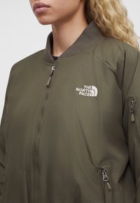 Γυναίκα με μπουφάν The North Face bomber σε ελιά πράσινο χρώμα με ριμπ υφασμάτινη λαιμόκοψη και τσέπες με φερμουάρ, εμφανιζόμενη από τον λαιμό έως τη μέση.