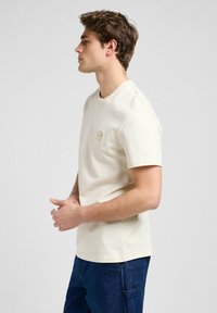 Creme kortærmet t-shirt med rund hals og en lille grafisk detalje på brystet, sammen med mørkeblå jeans. Glat tekstur.