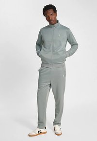 Lichtgrijs atletisch trainingspak van glad materiaal. Vest met ritssluiting, opstaande kraag en logo. Broek met elastische tailleband en taps toelopende pijpen.