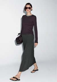 Vrouw met donkere zonnebril, paarse lange mouwen top, grijze geribbelde asymmetrische rok, zwarte sandalen, draagt een slordige donkere handtas, lopend.