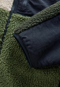 Fleecejakke i mørkegrøn og sort stof med tekstureret, blødt materiale og en glat ærme med lynlåsdetalje.