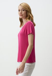 T-shirt fuchsia à manches courtes avec un décolleté en V, fabriqué à partir d'un tissu doux, associé à un pantalon blanc, vu de profil.