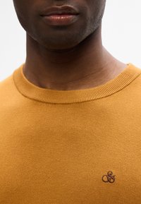 Musztardowy sweter z prążkowanym dekoltem i małym, ciemnobrązowym logo po lewej stronie klatki piersiowej. Gładka tekstura.