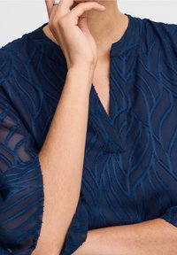 Laivansiniset paita, jossa on läpikuultavaa kangasta ja brodeerattu aalto-kuvio. Omistaa hienovaraisen V-kaulan ja röyhelömäiset hihat, jotka korostavat tekstuuria ja yksityiskohtia.