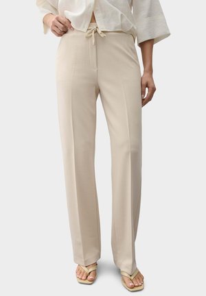 Person trägt beige weite Hosen mit Kordelzug in der Taille, weißes Oberteil und beige Flip-Flop-Sandalen, steht vor weißem Hintergrund.