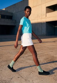 Jeune personne marchant à l'extérieur près d'un bâtiment en béton, portant un t-shirt bleu, un short blanc, des chaussettes vertes et des chaussures noires sans lacets.