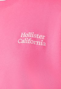 Rosa tyg med vit broderad text "Hollister California" nära bröstområdet på en klädesplagg.